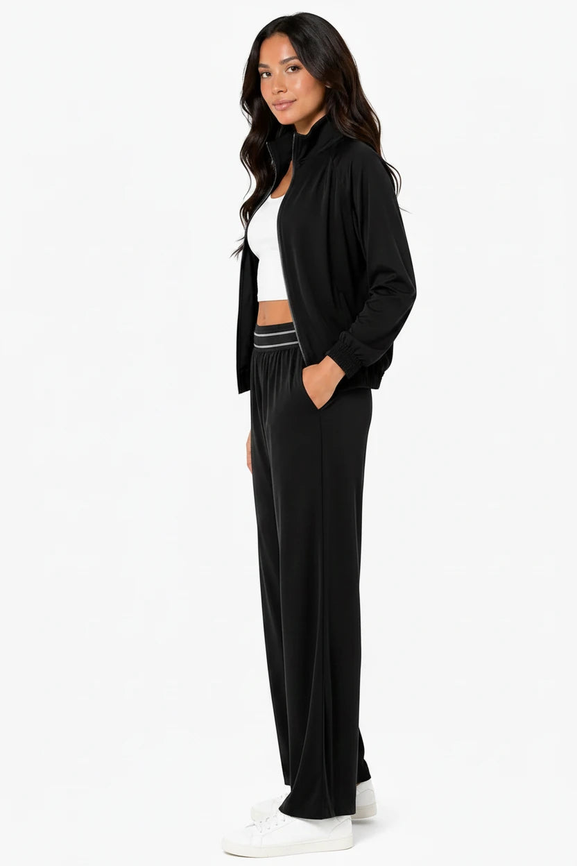 A woman modeling a black two-piece lounge set consisting of a lightweight zip-up jacket and matching wide-leg pants with a white crop top underneath, set against a plain white studio background.
