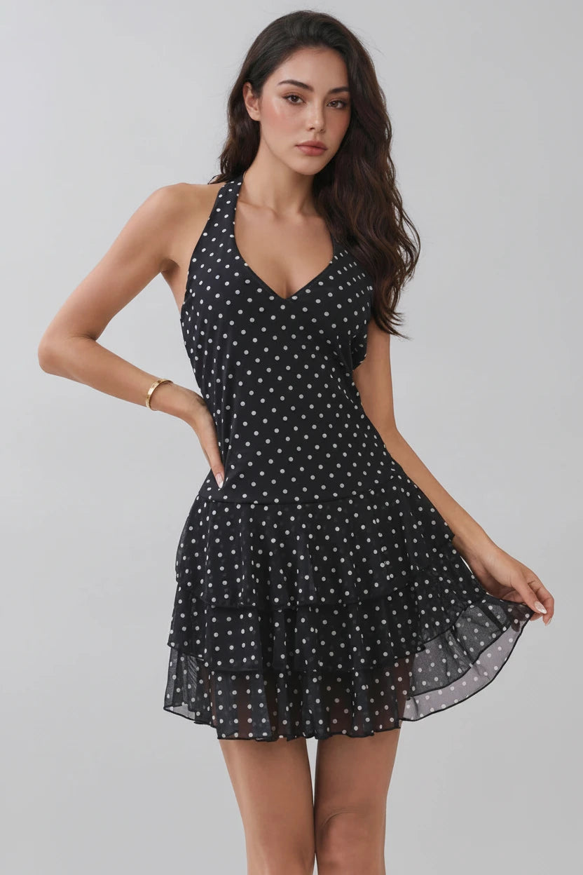 A woman modeling a sleeveless black halter mini dress with a white polka dot pattern against a light gray studio background.