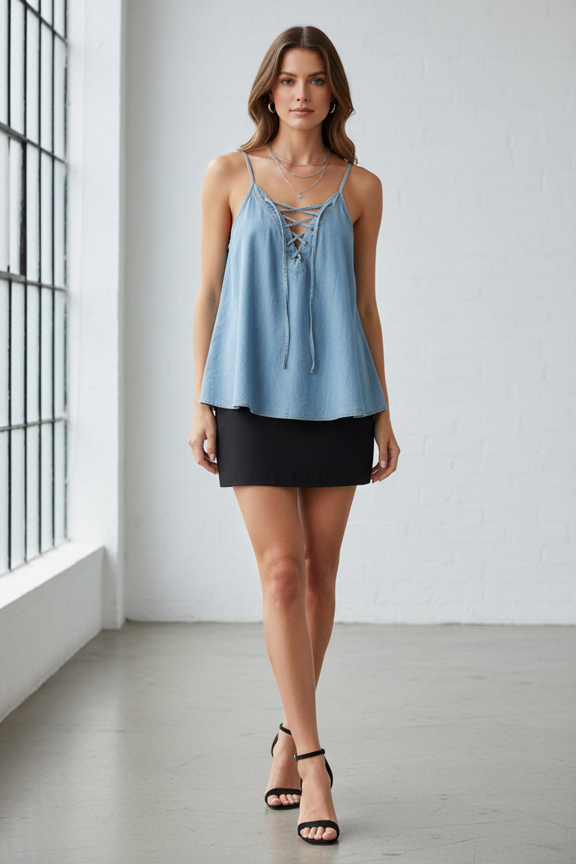 A blue jean blouse with lace up detail and adjustable straps, displayed on a hanger against a wooden background. It is paired with black pants.