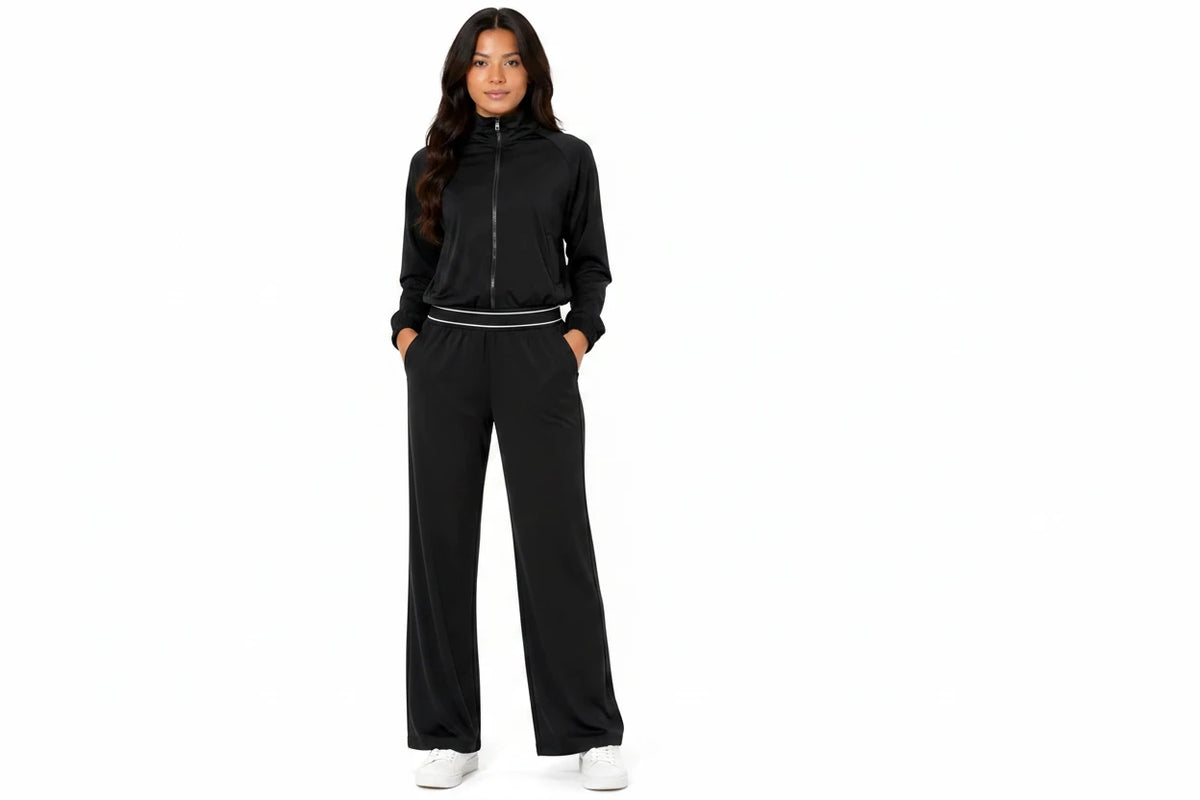 A woman modeling a black two-piece tracksuit with a zip-up jacket and matching wide-leg pants, featuring a thin white stripe detail at the waist, against a plain white background.