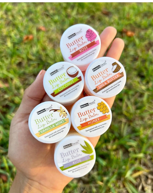 Hand holding six Beauty Treats Butter Lip Balms in assorted flavors" Rose,Coconut,Shea Butter,Vanilla Mint, and Aloe, displayed outdoors on grass.
